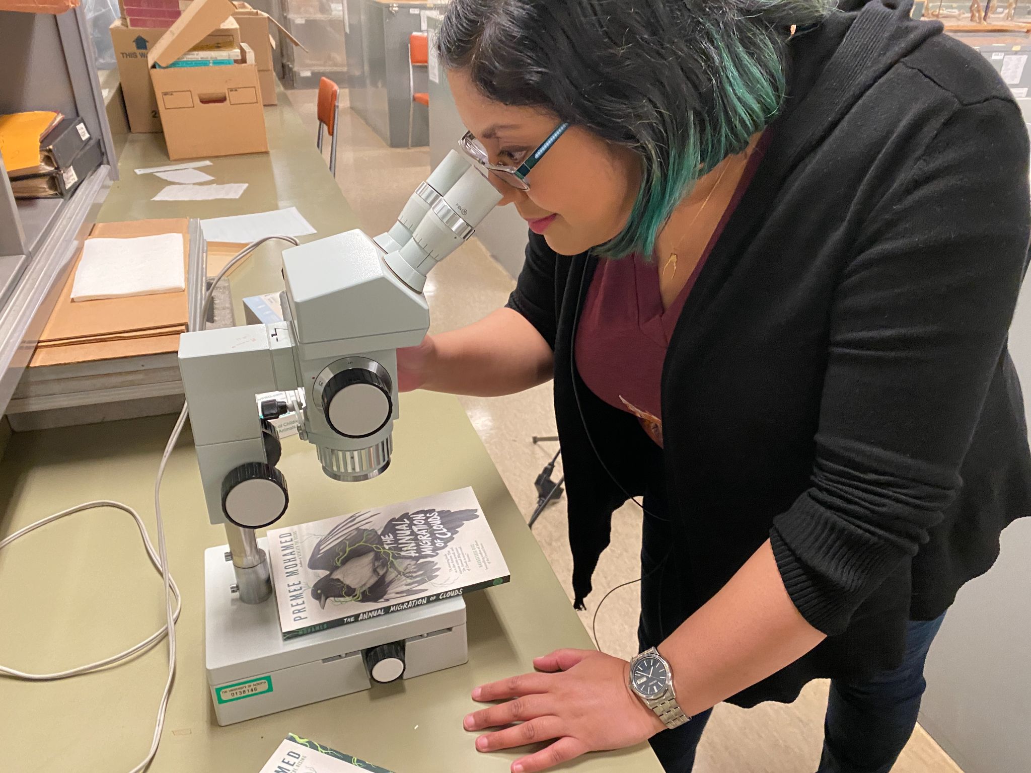 A woman with glasses looks at the front cover of the book, The Annual of Migrqtion of Clouds, through a microsope.