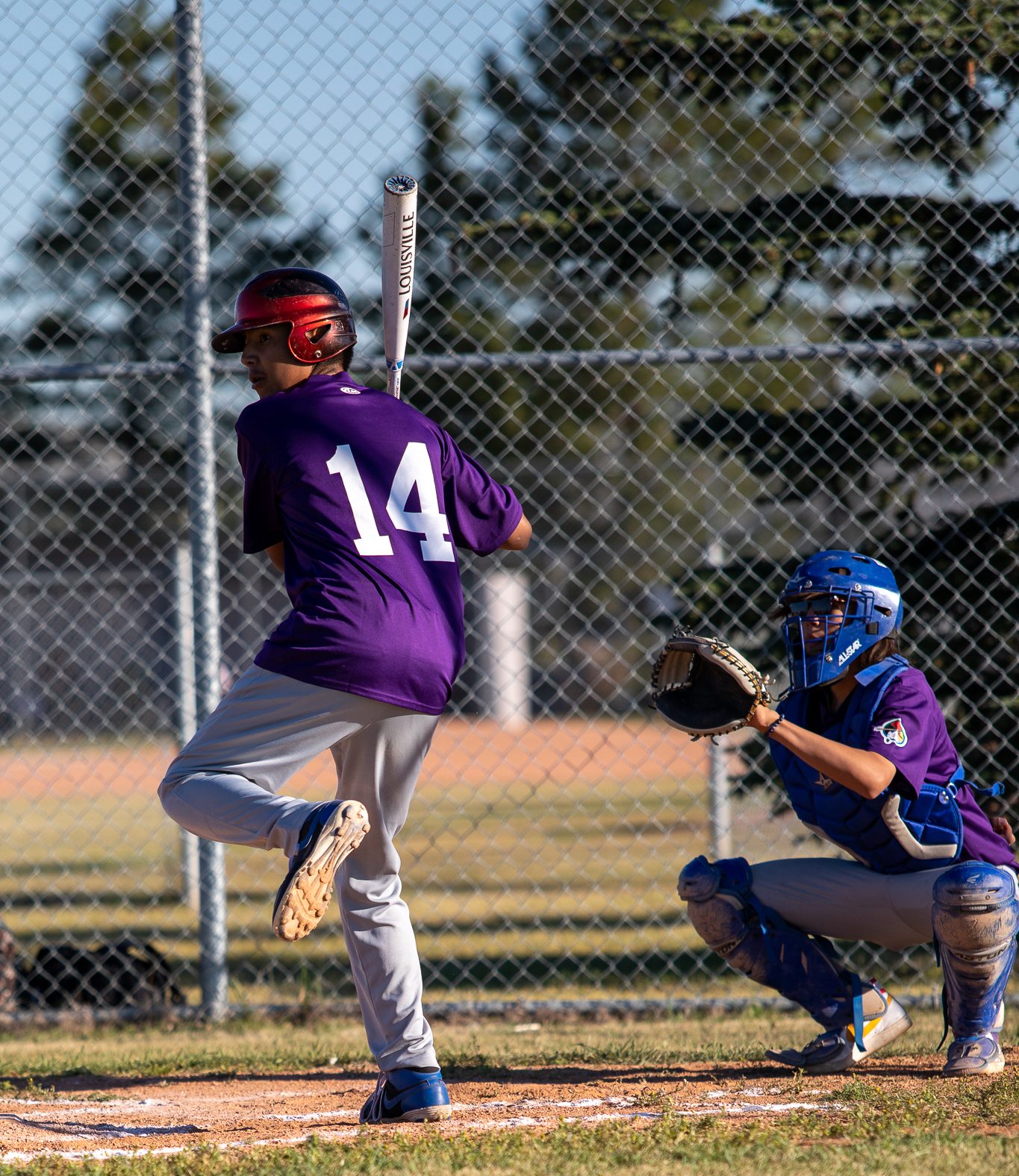 An young male Indigenous athlete winds up to swing a baseball bat as a catcher crouches behind him.