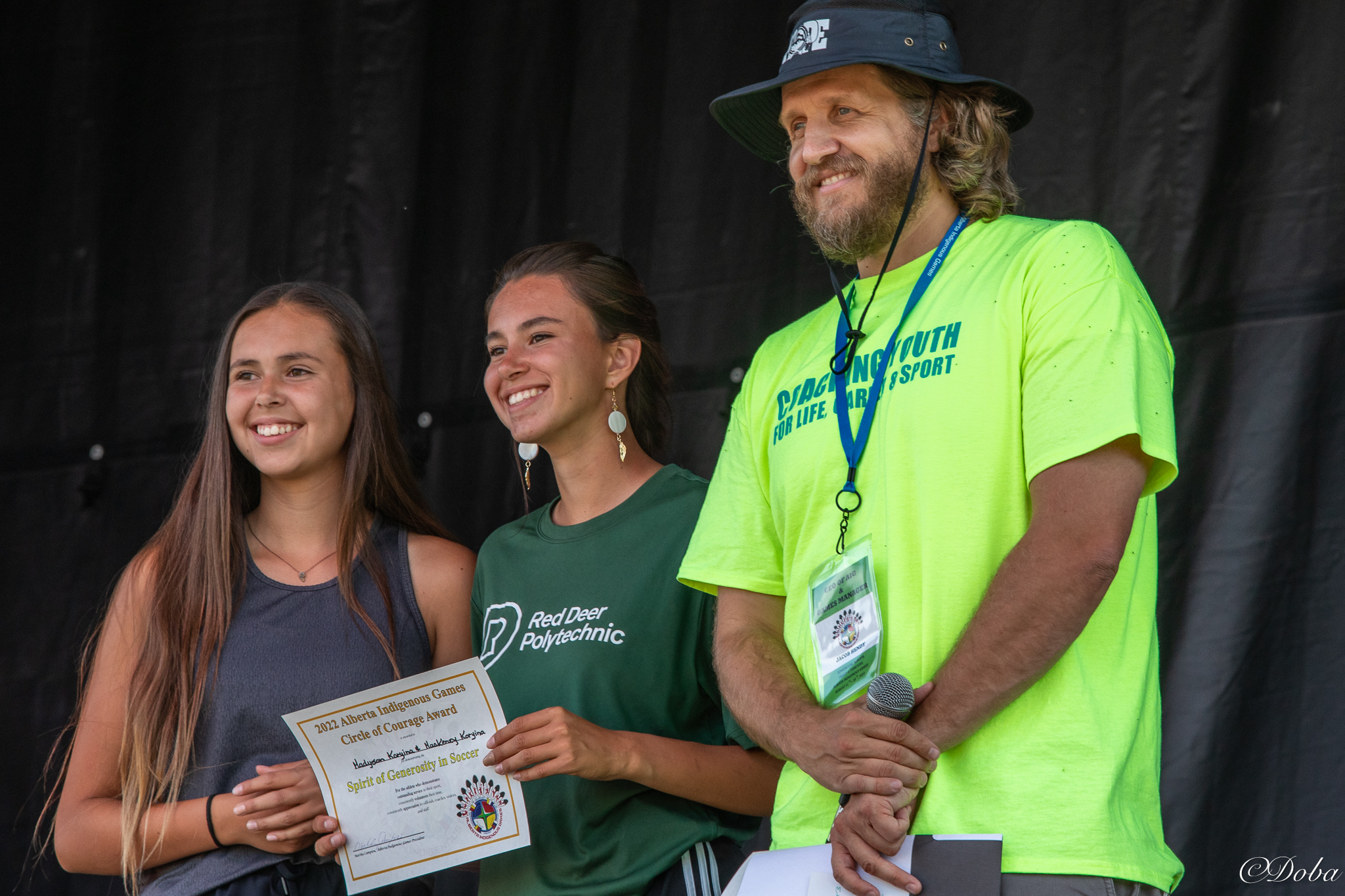 Two young Indigenous women stand next to a bearded man with a bucket hat.