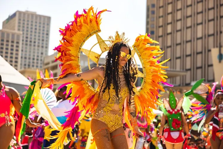 Một nghệ sĩ biểu diễn tại Lễ hội Cariwest trong bộ trang phục màu vàng rực rỡ được trang trí bằng lông vũ màu vàng và hồng đang nhảy múa tại một quảng trường công cộng đông người.