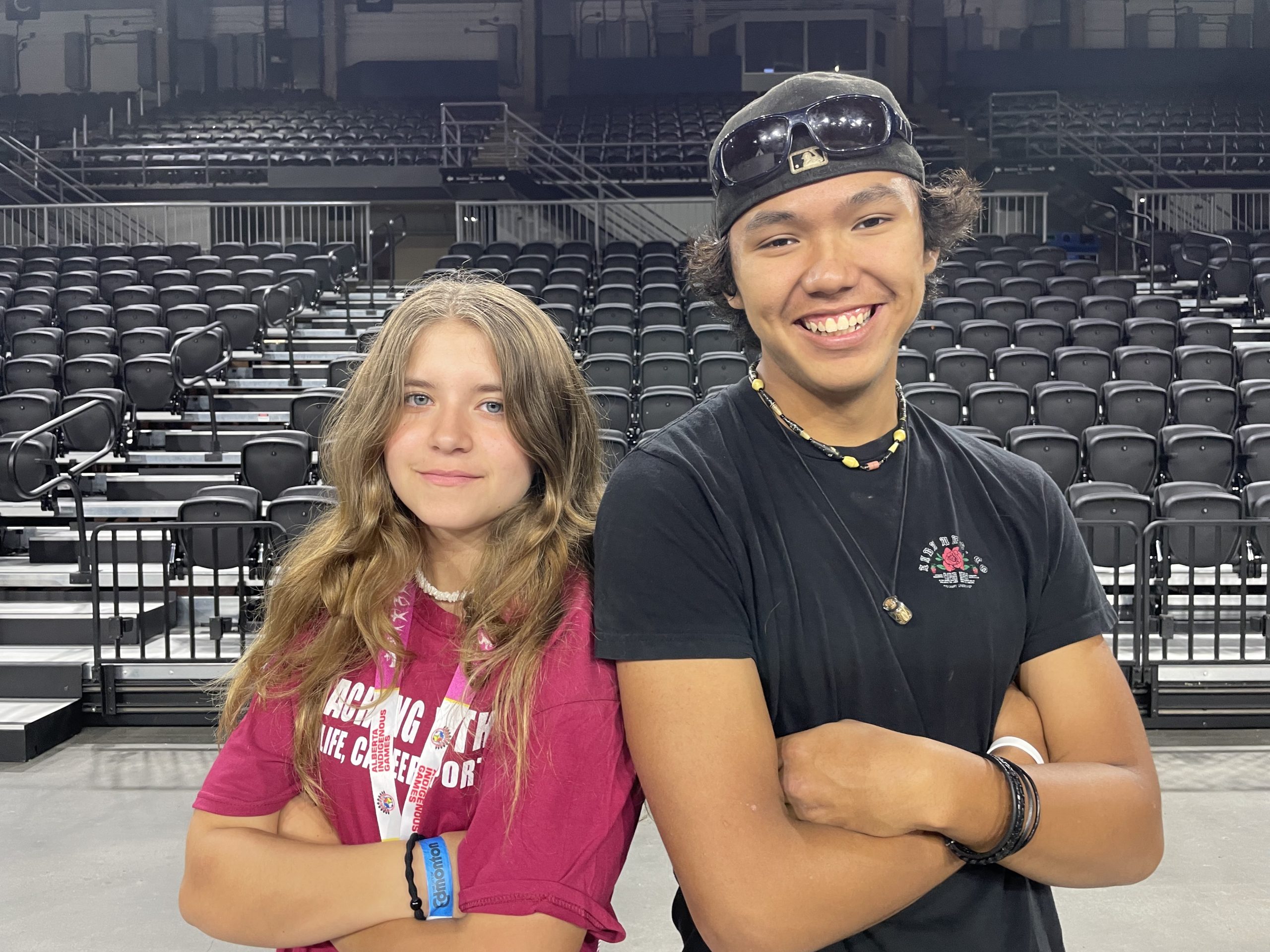 Two teens, a girl and a boy, stand next to each other with their arms crossed.