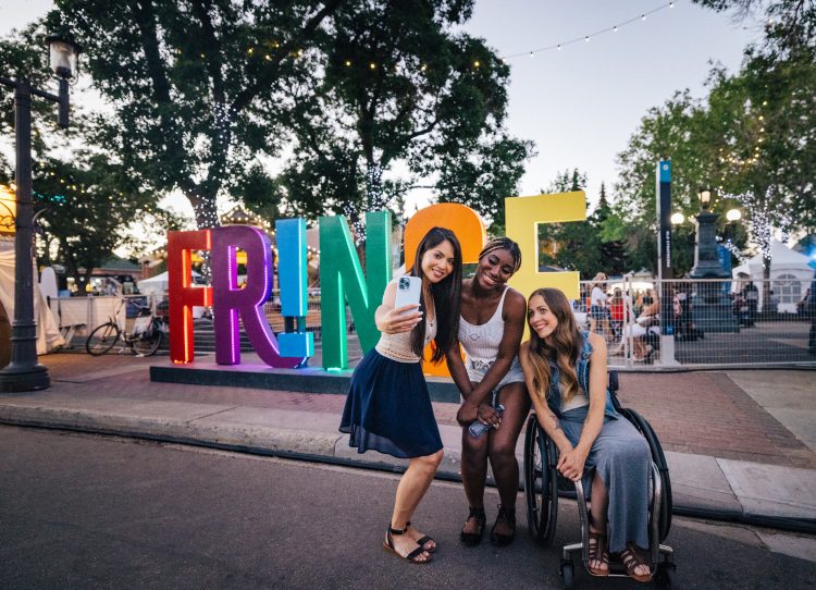 Three women stand in front an art installation with the words "FRINGE" in large colourful lettering, taking a selfie