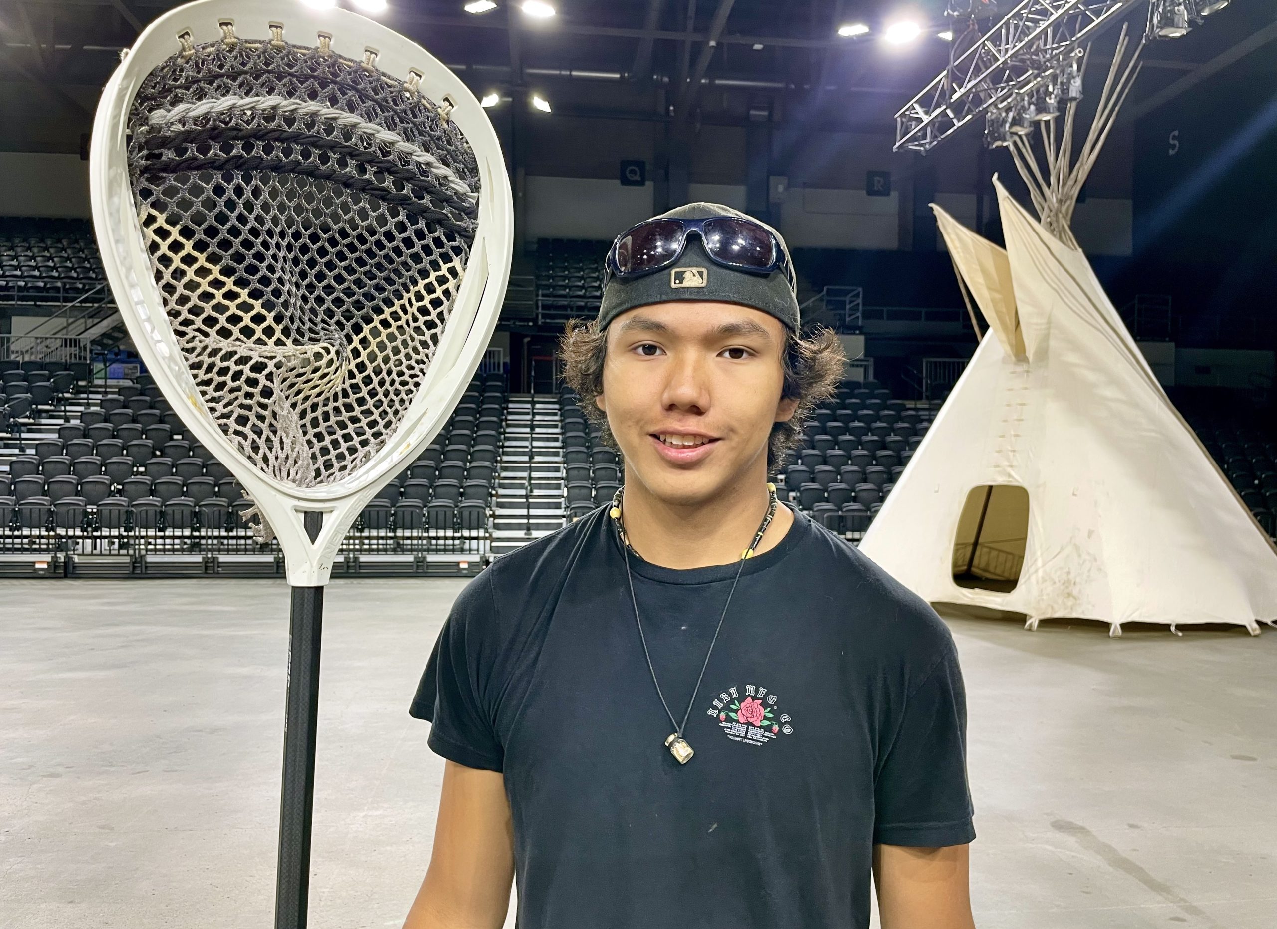 A young male Indigenous teen, holding a lacrosse stick, stands in front of a tipi inside a small basketball arena.