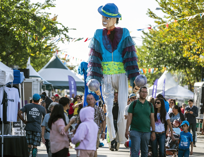 A giant character on stilts walks down a crowded festival street,