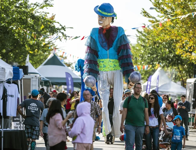 A giant character on stilts walks down a crowded festival street,
