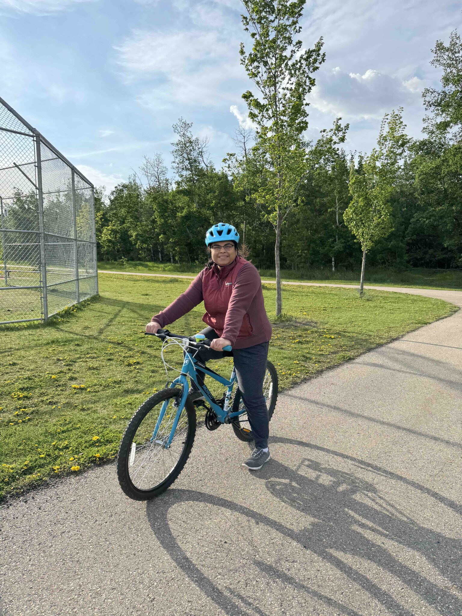 A woman on a blue bike smiles at the camera on a shared use pathway near a park.