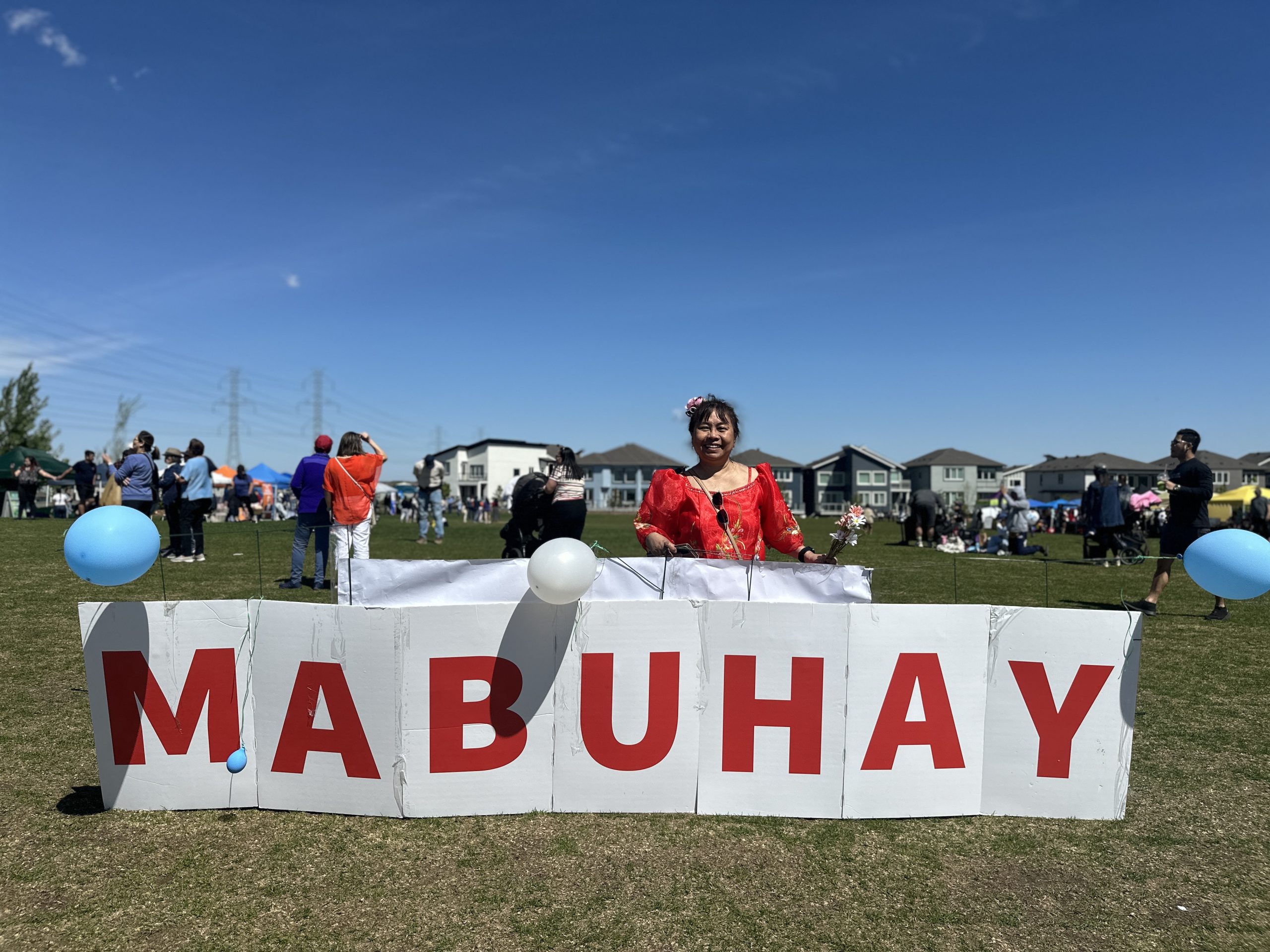 A woman stands behind a sign that reads Mabuhay in the middle of a park lined with homes.