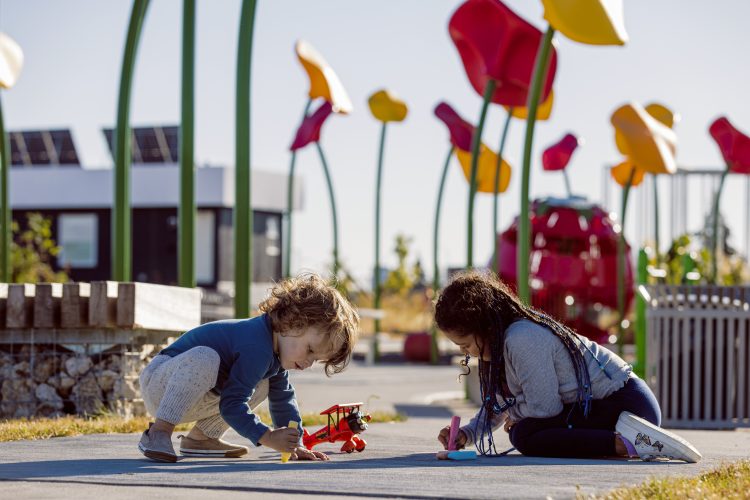 Two children draw with sidewalk chalk in a park with large floral art installation in the background