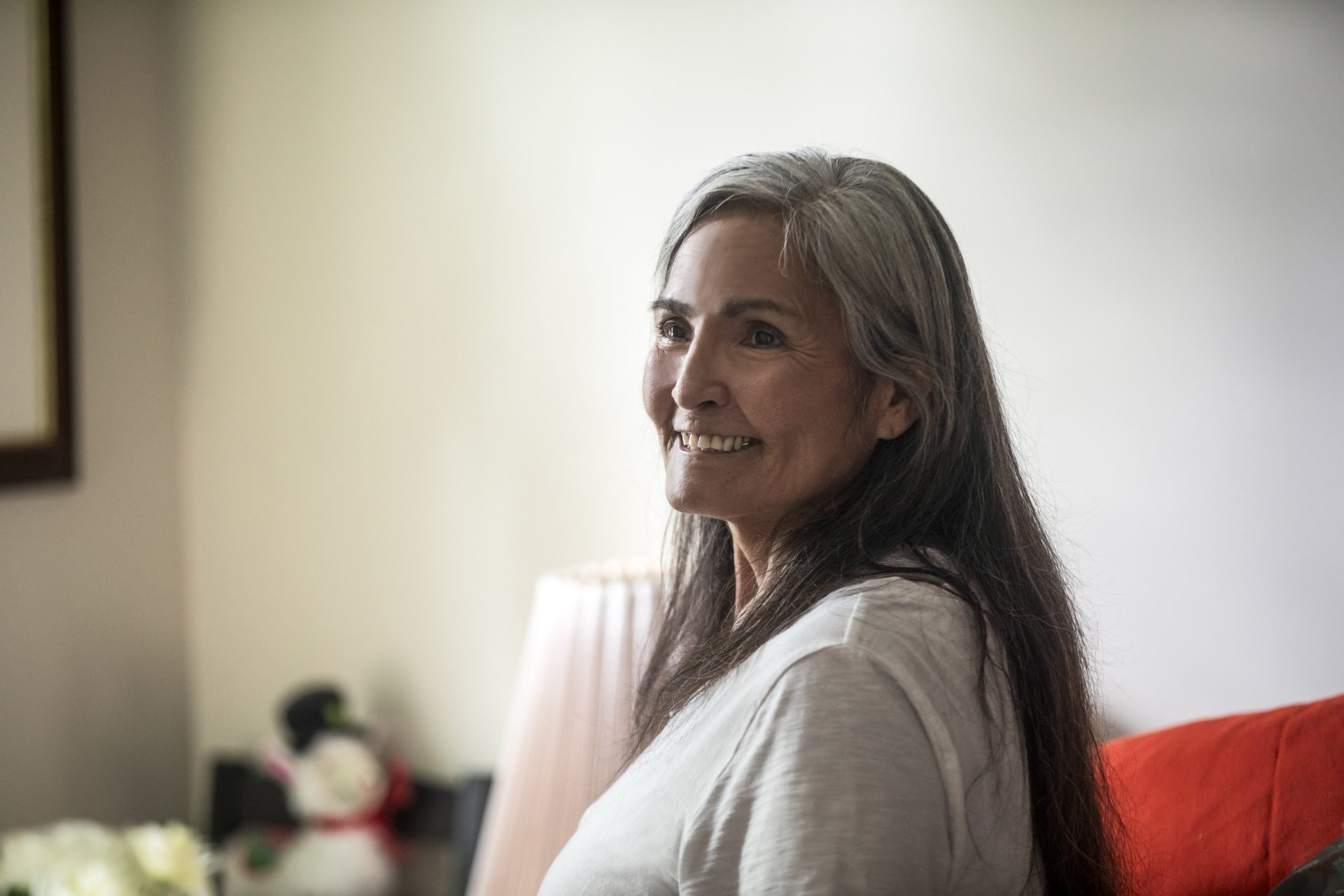 An Indigenous woman with long brown and grey hair smiles as she sits in her home.