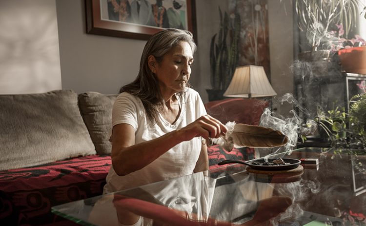 An Indigenous woman fans an eagle feather over a smudging pot of sweetgrass as she sits on the floor between her couch and coffee table.