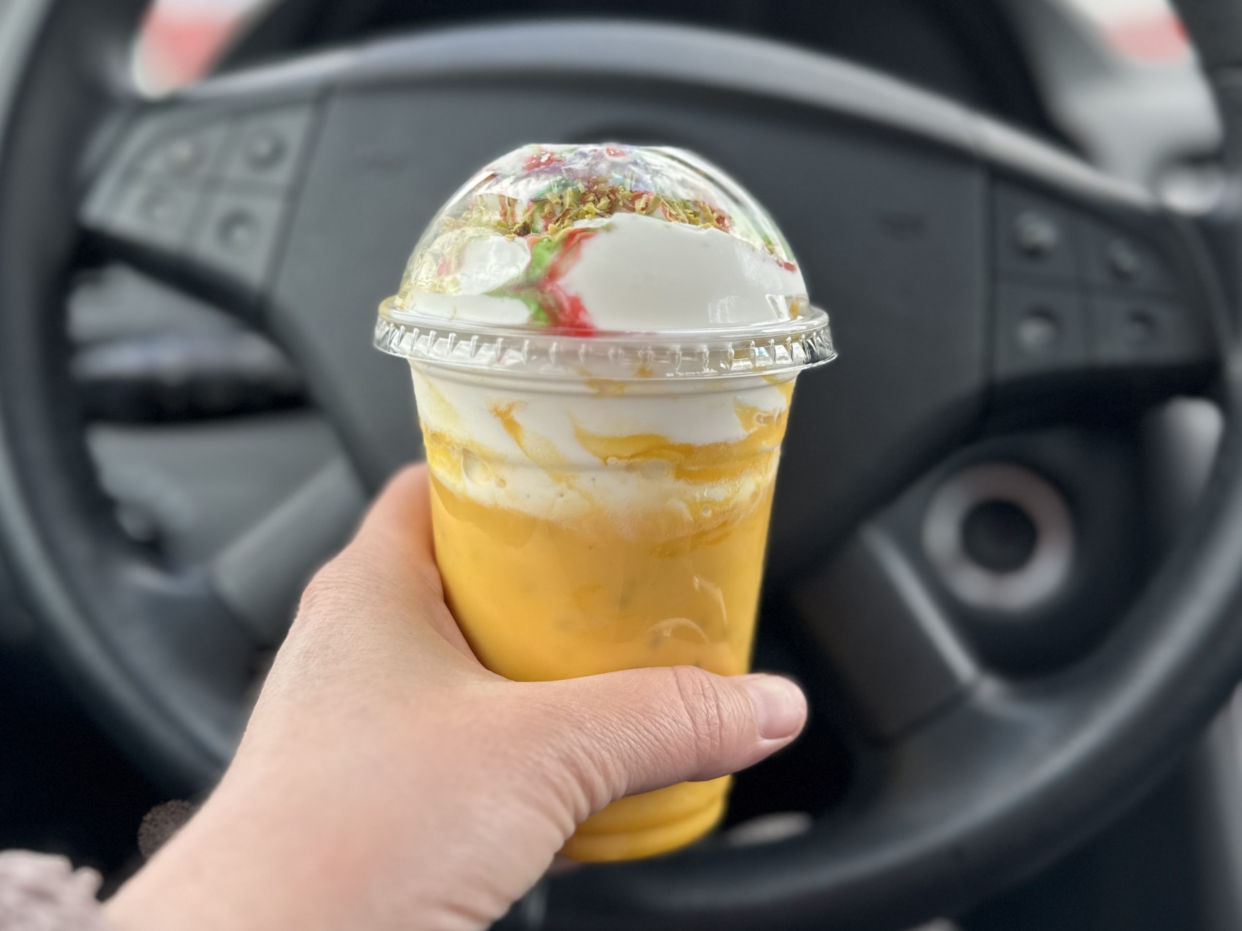 A hand holds an ice cream drink in front of a steering wheel of a car.