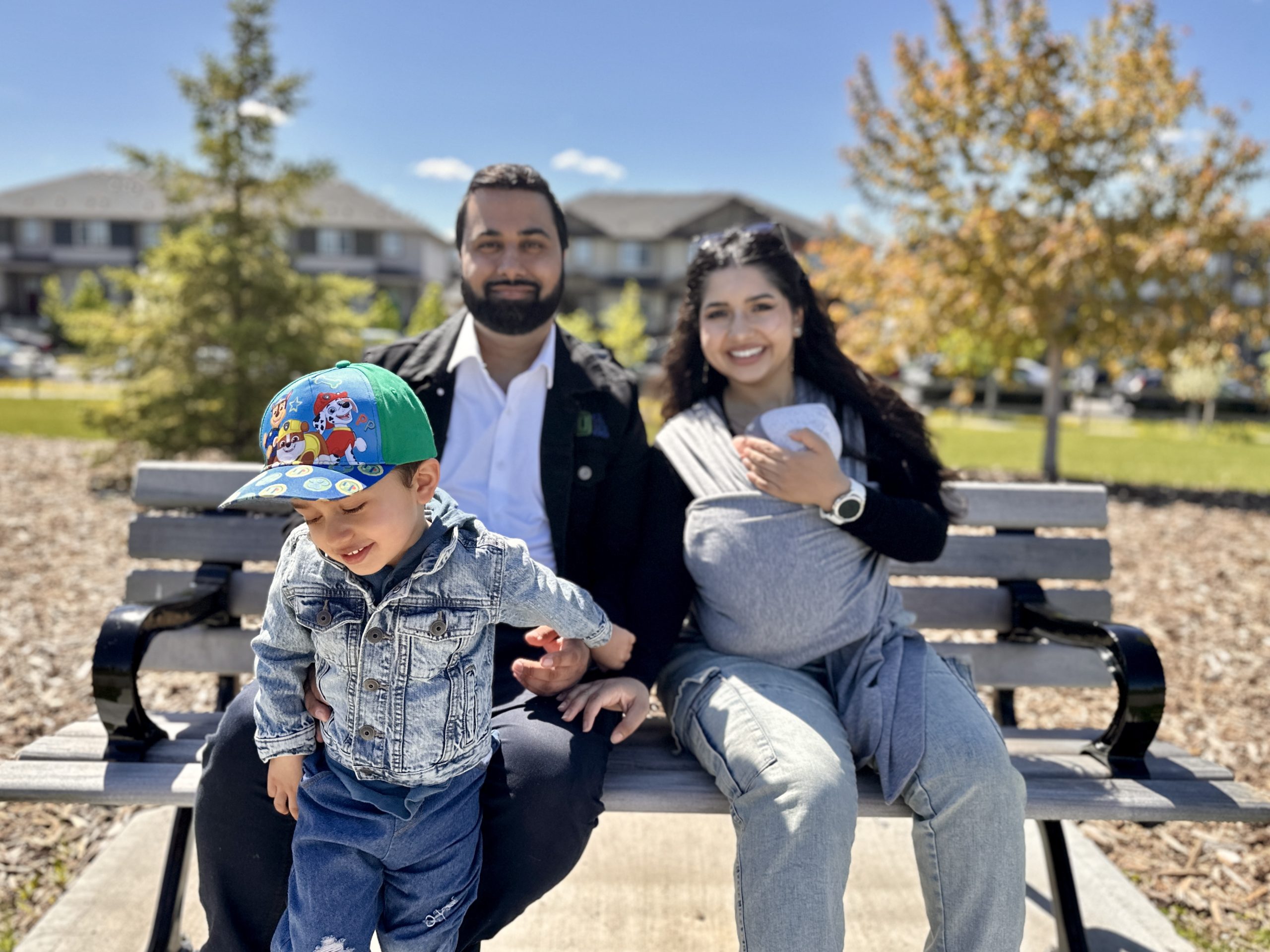 A husband and wife, with a baby in a sling, sit on a park bench as their young son stands in front of them.