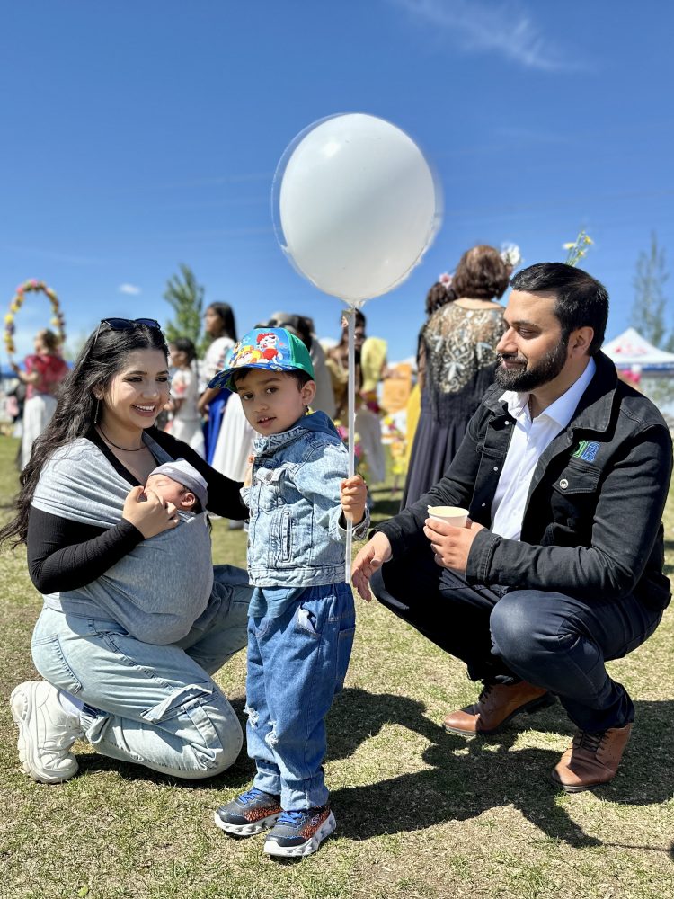 A boy, holding a balloon, stands between his kneeling father and mother, who is carrying a baby in a sling.