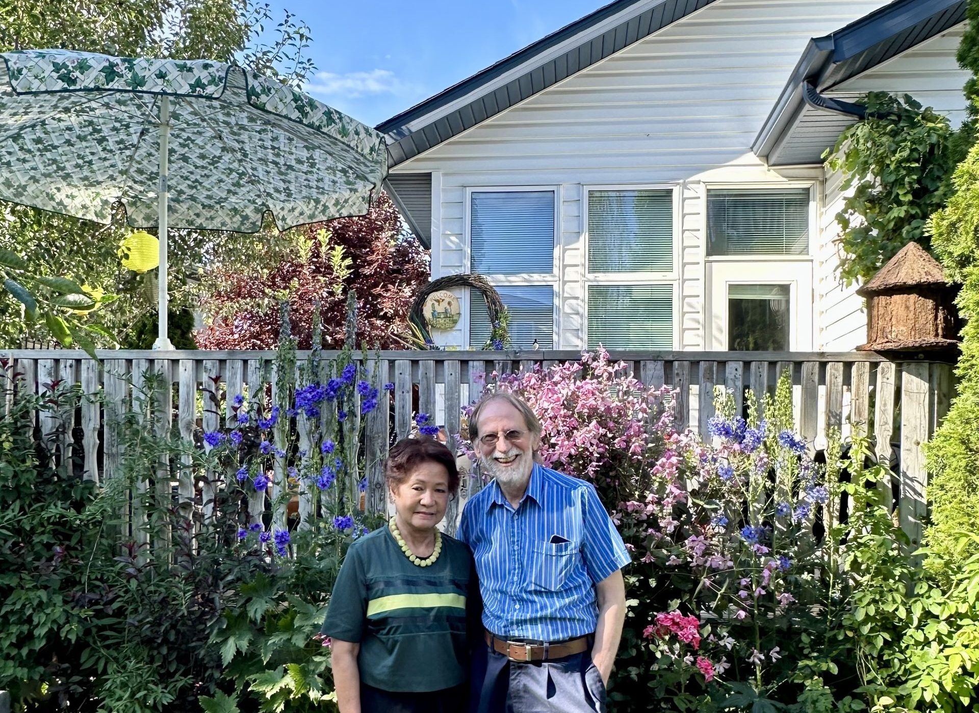 A woman and man stand in front of their back deck, surrounded by flowers and trees.