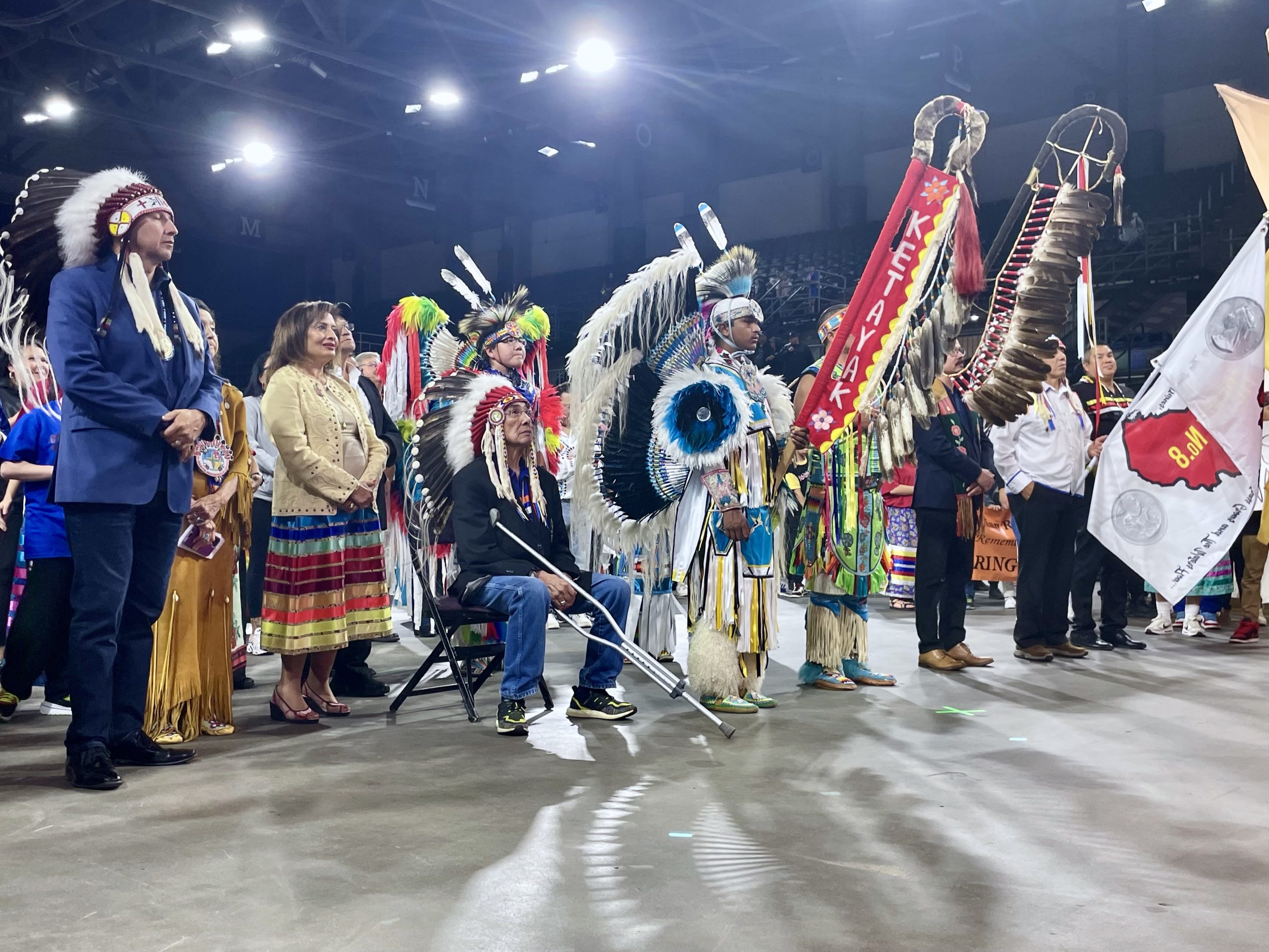 A group of Indigenous women and men, some in traditional head dresses and outfits, stand and sit on the floor of a basketball arena.