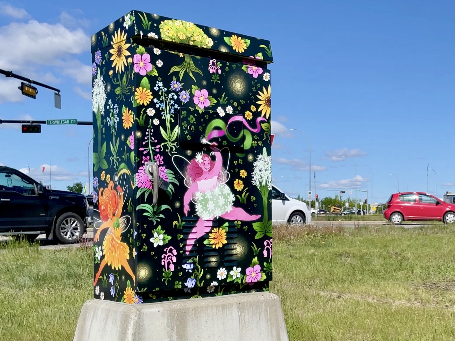 Cars drive by a traffic control box wrapped with images of neon fairies and flowers.