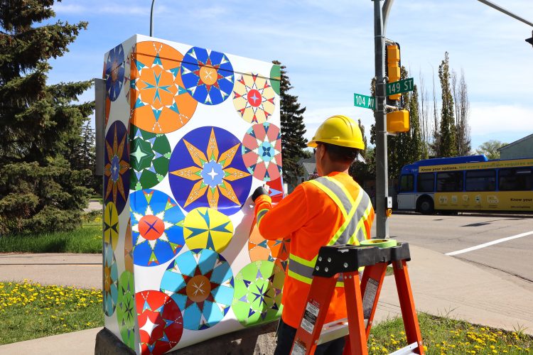 A City of Edmonton worker in an orange safety shirt wraps a traffic control box with artwork featuring kaleidoscopic circles.