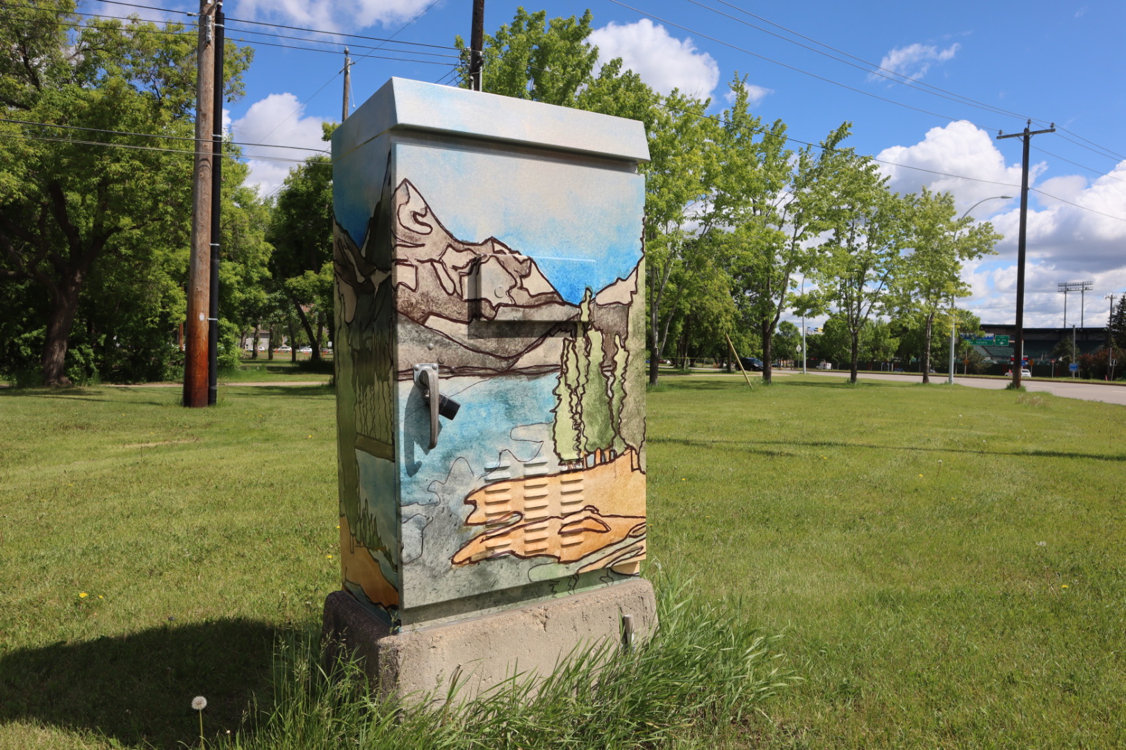 A traffic control box, standing in a grassy field, is wrapped with a painting of mountains and a tree-covered island in a lake.