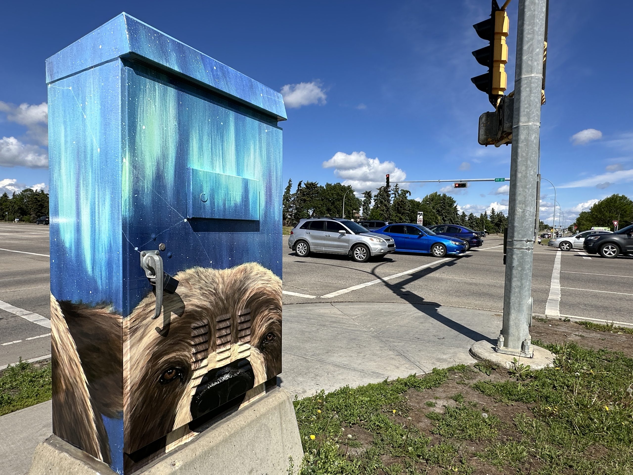 A painting of a bear's face peeks out from the bottom of a traffic control box as cars drive by at an intersection.