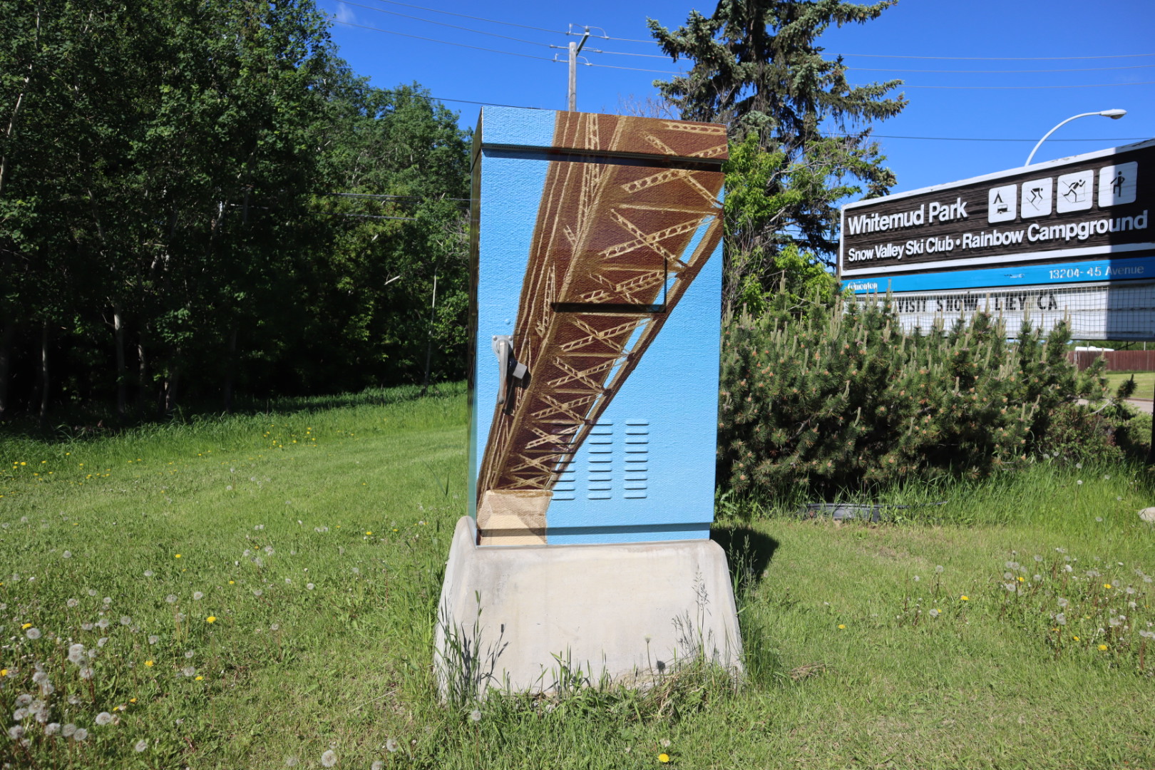 A traffic control box wrapped with artwork of the High Level Bridge stands in a grassy field next to a Whitemud Park sign.