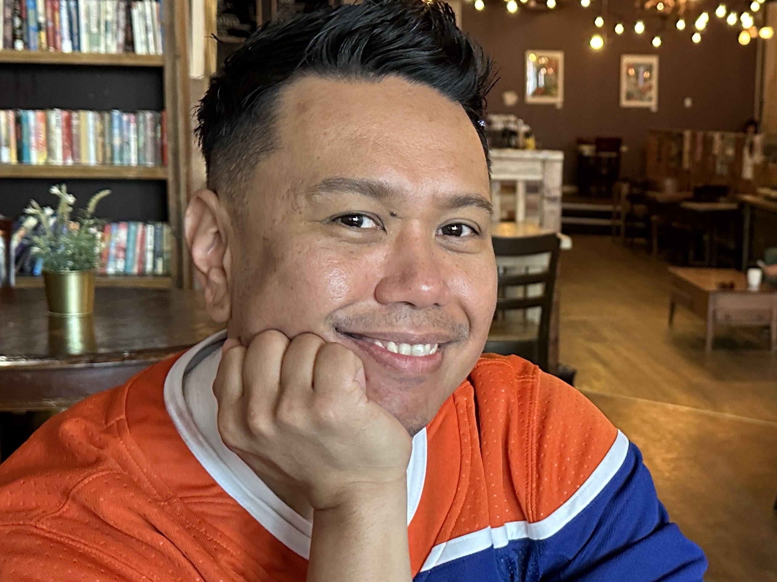 A man, wearing an Oilers jersey, smiles as he rests his chin in his hand in a café.