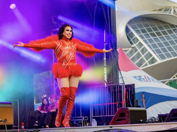 A drag queen in a red bodysuit raises her arms to the side on a stage next to a tent and a wavy metallic building.