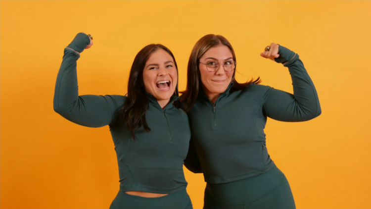 Edmonton’s Amazing Race Canada team, Taylor McPherson and Katie Mulkay. Two brunette women standing flexing their arm muscles and laughing.