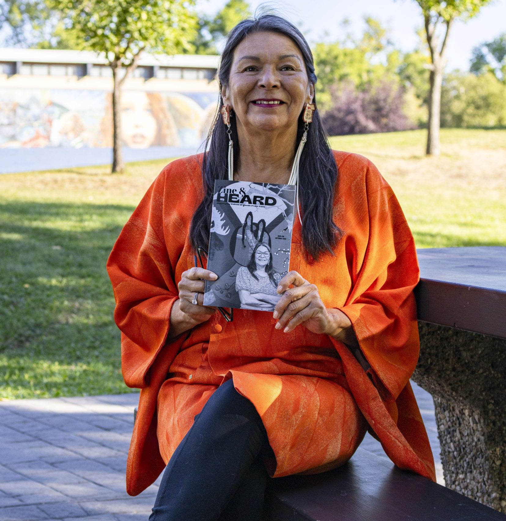 A smiling Indigenous woman holds up a copy of a zine as she sits at a picnic table in a park.