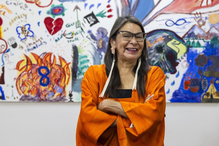 A smiling Indigenous woman wearing an orange dress stands in front of a painting.