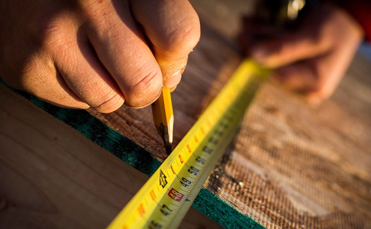 Close up of a man’s hands using a pencil to mark wood with a measuring tape.