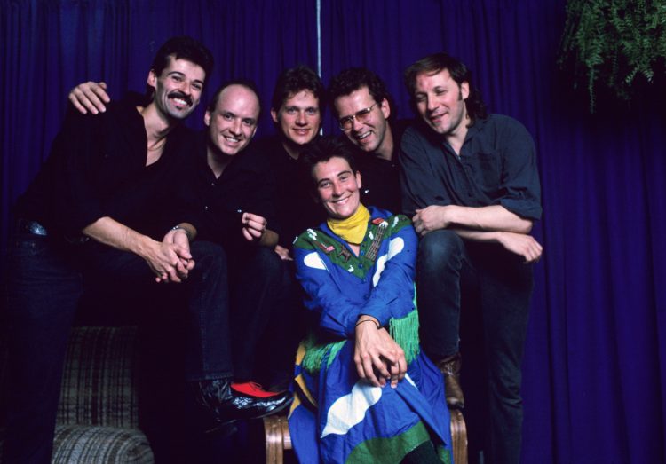 Five men in jeans and long-sleeved shirts stand around a woman in a blue, green and white western dress sitting in a chair.