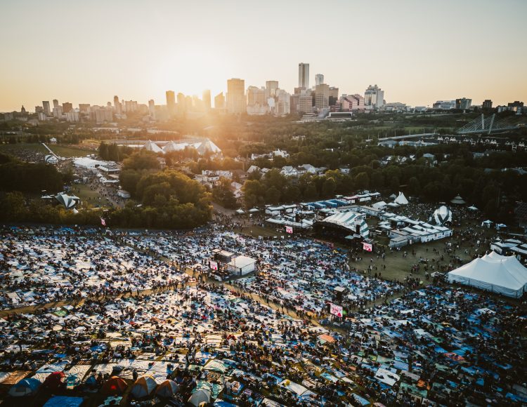 Edmonton Folk Fest hill filled with people watching performances on the stage. The sun is setting behind the skyline in the background with a ray of light.