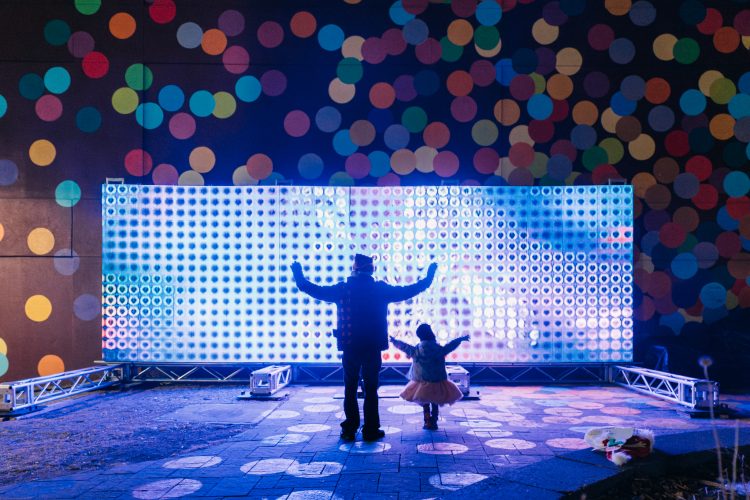 An adult and child standing with their arms out are illuminated by a giant light screen behind them at an outdoor winter festival.