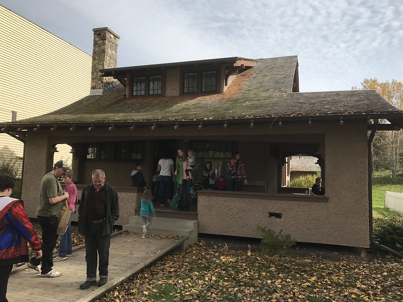 Guests exploring the exterior of a small historic house with a wrap around porch, half second story and steep roof on a sunny fall day.