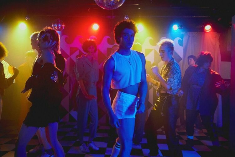 A 80s nightclub featuring a man dancing in white cutoff jeans and mesh crop top in colourful strobe lights.