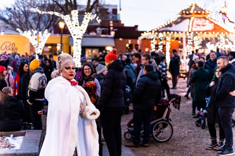 A woman in a white coat featured in a crowd enjoying an outdoor winter festival with many twinkling lights.