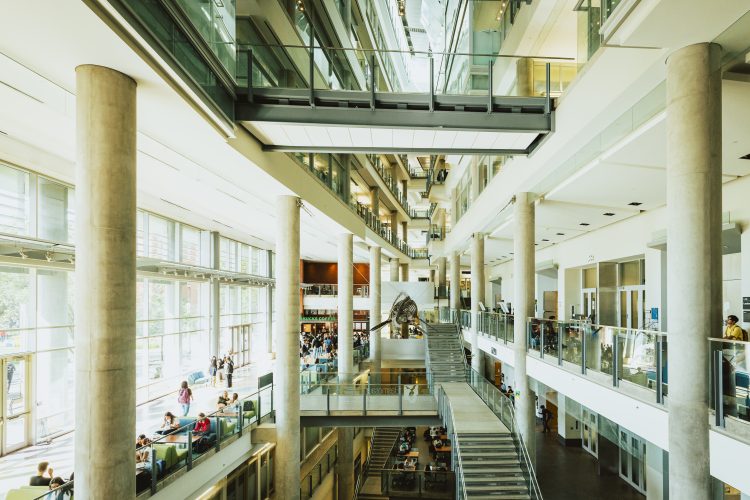 Two-story atrium inside a post secondary building with stairs, glass and concrete pillars.