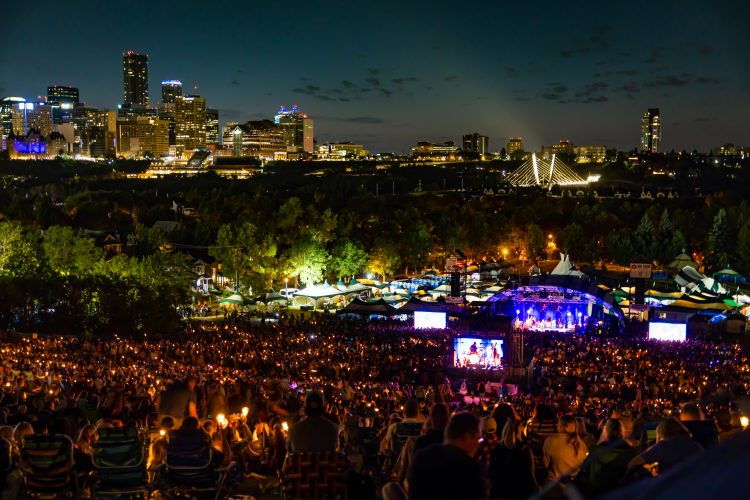 Wide shot of a night crowd sitting down Gallagher Park hill watching a performance during the 2024 Edmonton Folk Festival, with the city skyline illuminated in the background.