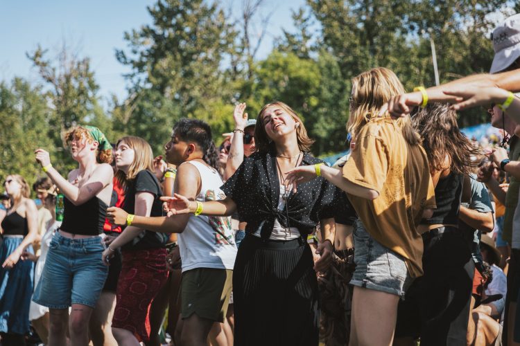 Festival goers dancing to live music with trees and sunlight in the background at the Edmonton Folk Festival.