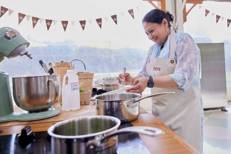 A woman in an apron works at a kitchen counter with a stand mixer and pots on a stove.