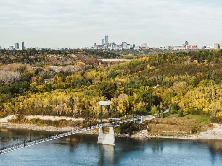 A footbridge across a river in a fall valley with the city skyline in the background.