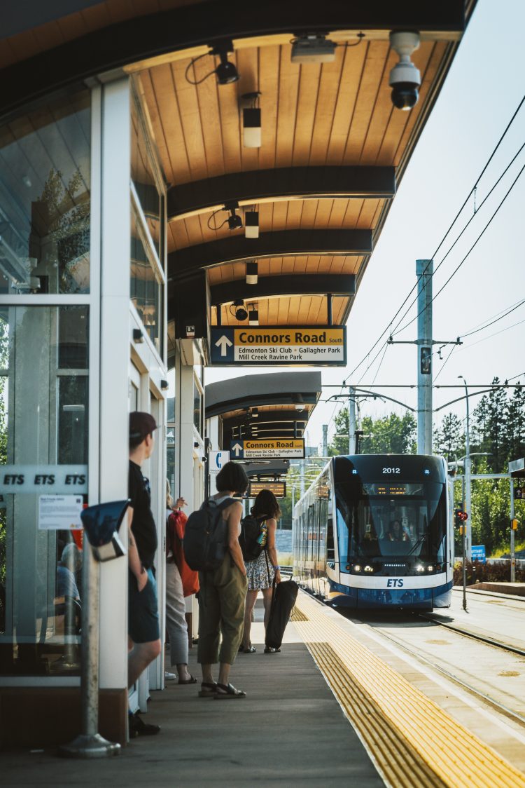 The L-R-T arriving at the Edmonton Muttart L-R-T Station on a sunny day. People are waiting on the platform and directional signage above indicated Connors Road.