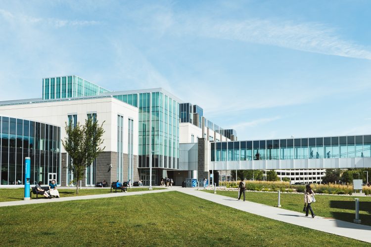 A glass pedway attached to a modern 4-story building with blue glass windows and concrete blocks.