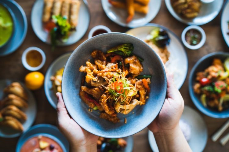 Overhead shot of many Asian dishes on a table with a noodle bowl being held up with 2 hands.