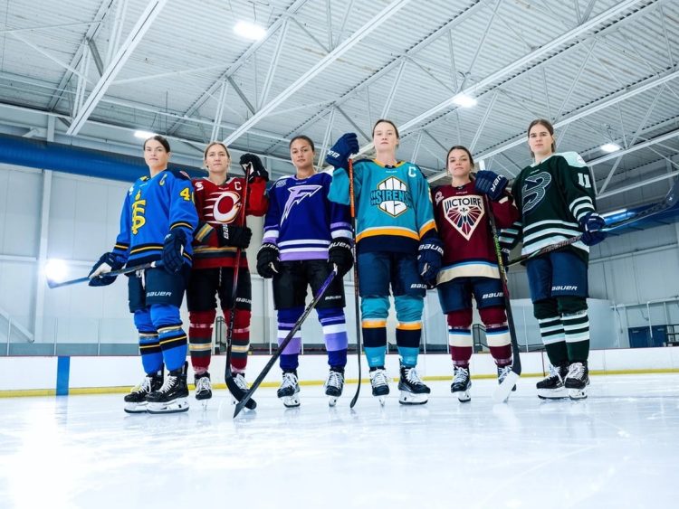 Six female professional hockey players in different uniforms standing on an indoor ice rink.