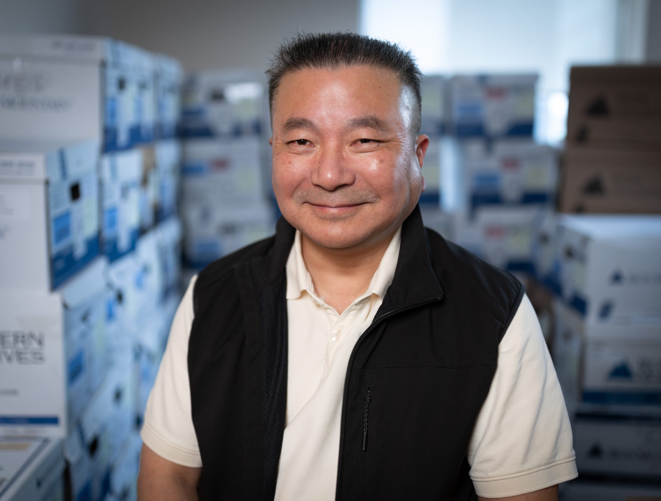 A man smiles as he stands in a room filled with cardboard boxes with lids.