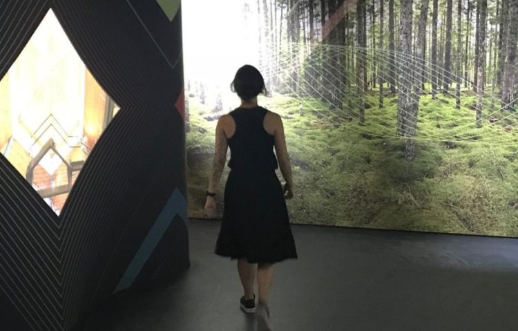 The back of a woman wearing a black dress is walking through an exhibit featuring a fill wall with an image of a forest.