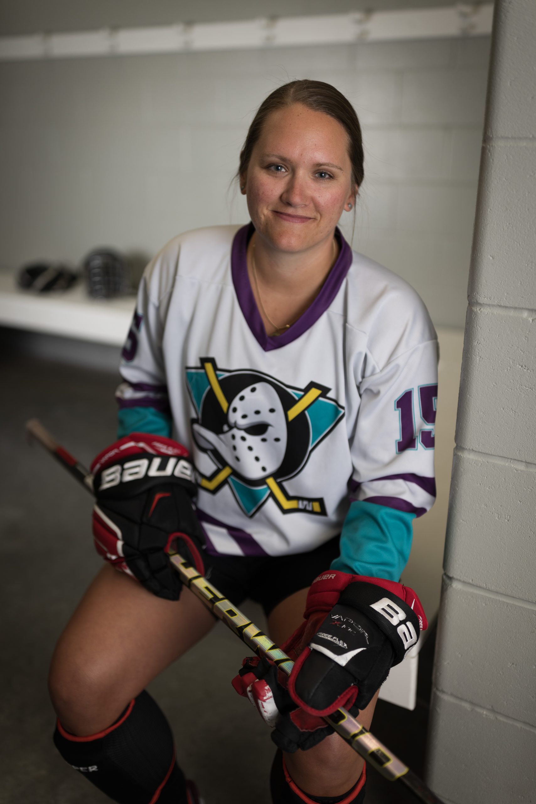 A woman in a ball hockey uniform, including a white jersey with a duck logo, gloves and a stick sits in a dressing room.