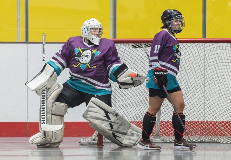 Two women playing in a ball hockey game; the goalie and a forward, both wearing purple jerseys with ducks crests.