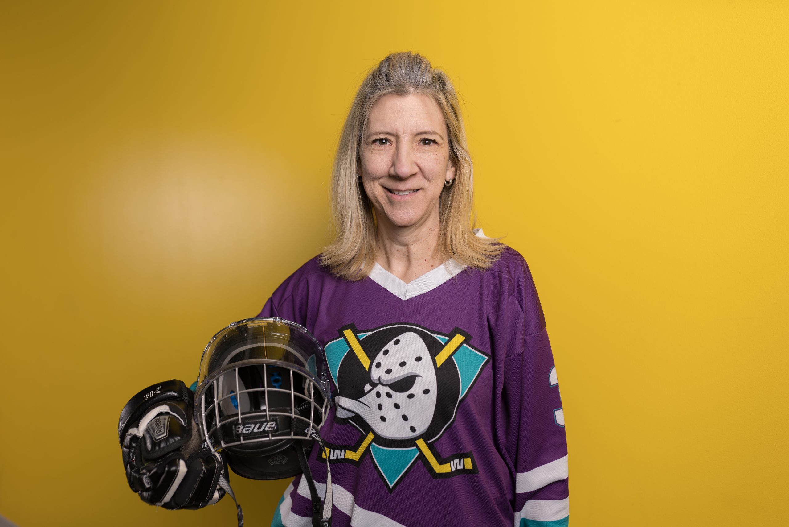 A woman wearing a hockey jersey and holding a helmet smiling in front of a yellow wall.